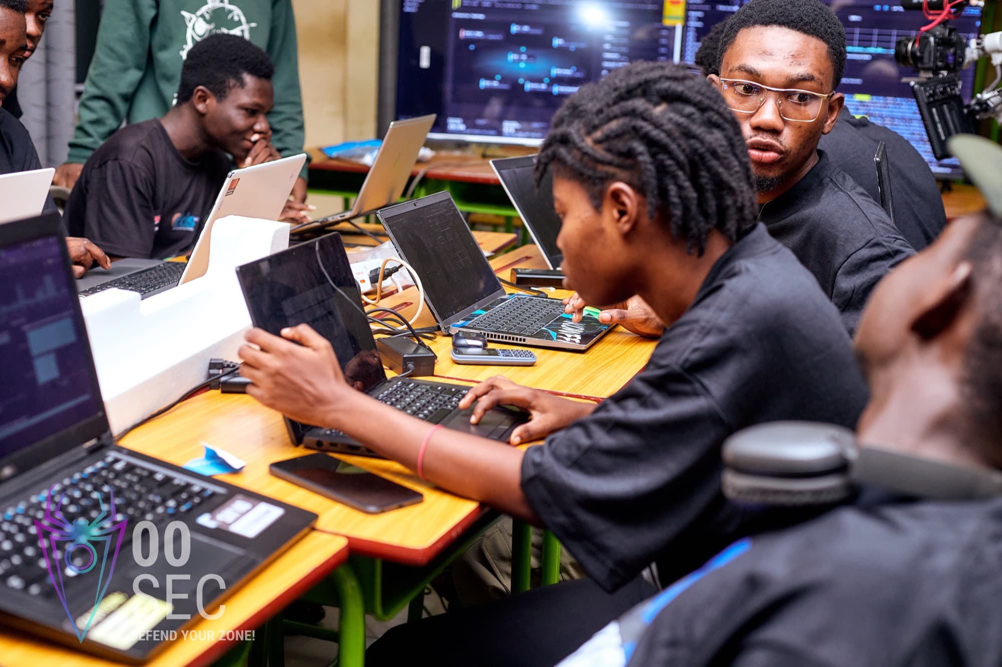 Participants deep in a CTF challenge on their laptops, live scoreboard data on the screen behind them.